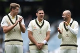 Two cricketers clap and look at a dark haired cricketer between them in middle of a big stadium