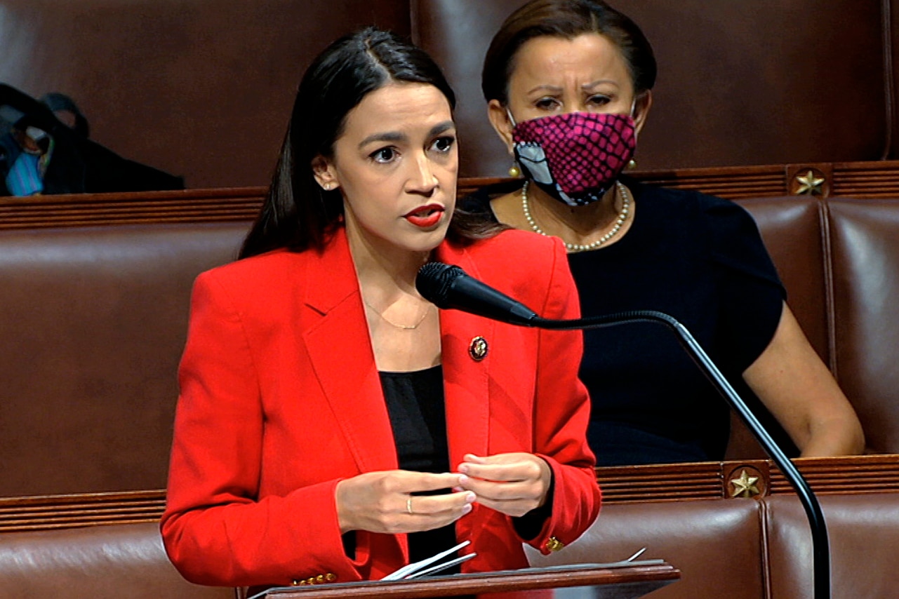 Alexandria Ocasio-Cortez speaks into a microphone she is wearing a red blazer