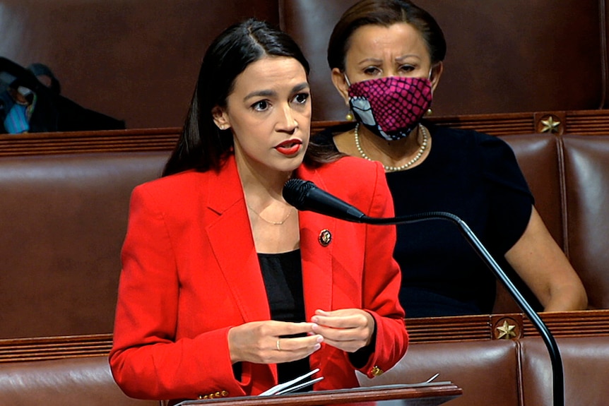 Alexandria Ocasio-Cortez speaks into a microphone she is wearing a red blazer