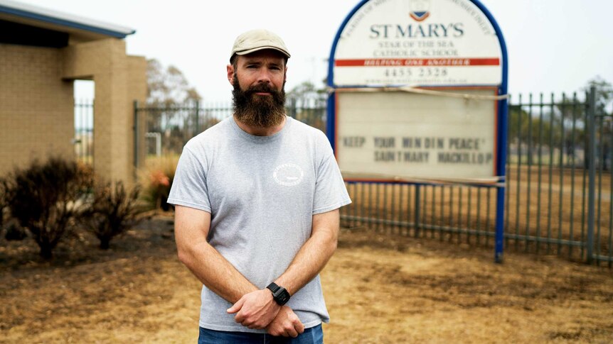 A bearded teacher stands in front of a school sign
