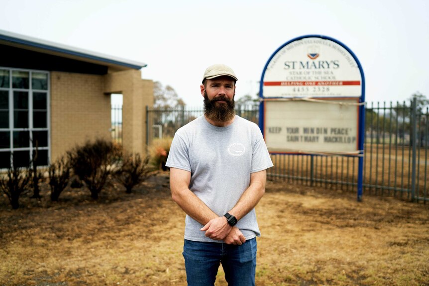 A bearded teacher stands in front of a school sign