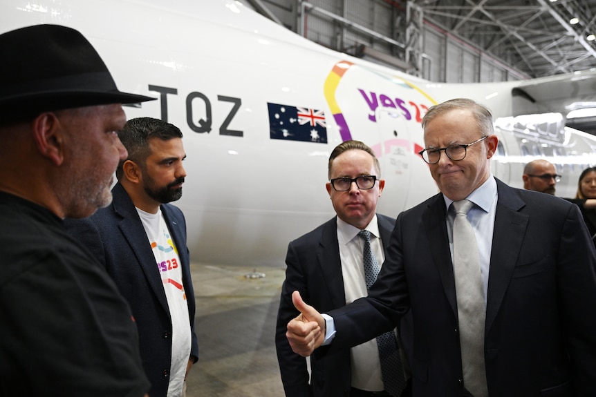 Four men wearing suits stand next to a Qantas plane with the Yes campaign logo on its side. 