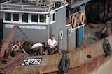 Looking down at a grey and rusting fishing boat with three men on deck watching their fishing roads to the side of its deck.