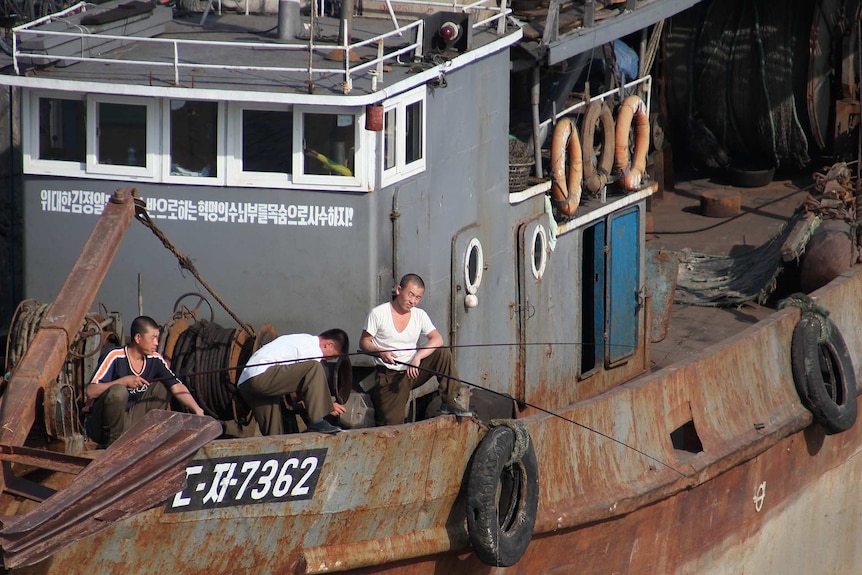 Looking down at a grey and rusting fishing boat with three men on deck watching their fishing roads to the side of its deck.
