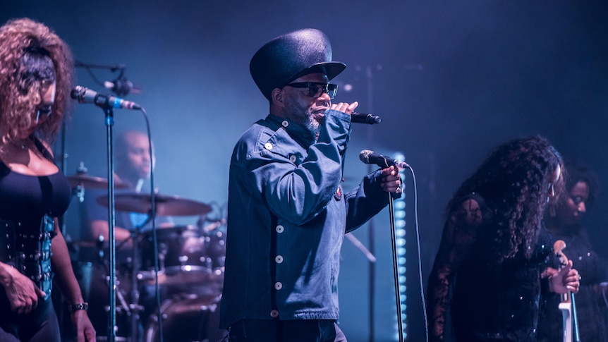 three people stand on a dark stage, singing into microphones, Jazzy B, a man in the middle wears a large hat