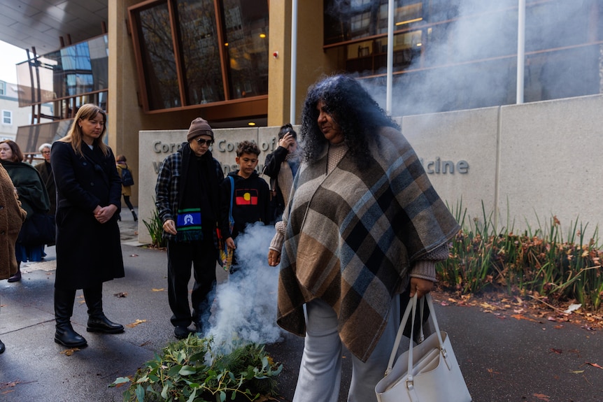 Donna Kerr wears a brown checked shawl and walks through the smoke from burning gum leaves outside court.