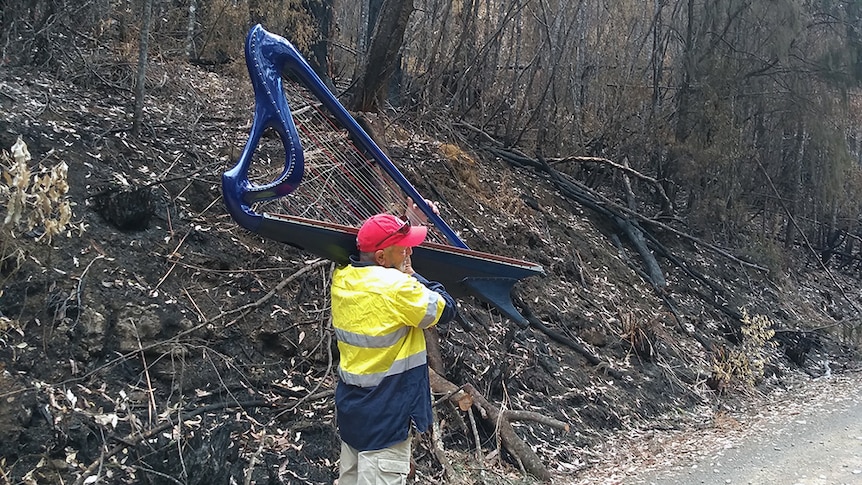 A man in a yellow high-vis jacket carried a blue harp through bushfire-damaged landscape in Tasmania.
