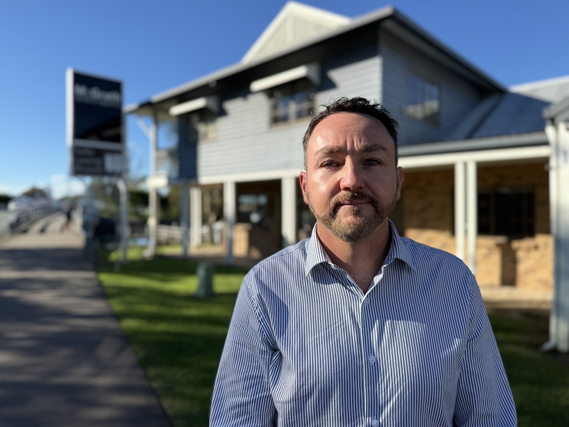 A man with short dark hair, wearing a business shirt, standing outside a house.