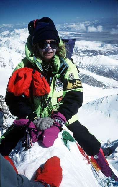 Brigitte Muir with white zinc or her face, sunglasses, beanie and snow clothing, on a snow-capped peak with mountain surrounds.