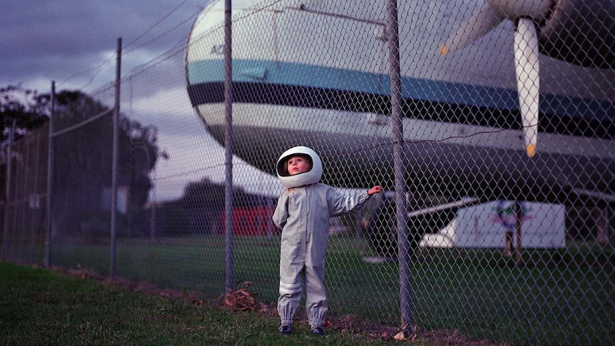 A young girl wearing a spacesuit costume and helmet standing in front of a wire fence and a plane with propellers