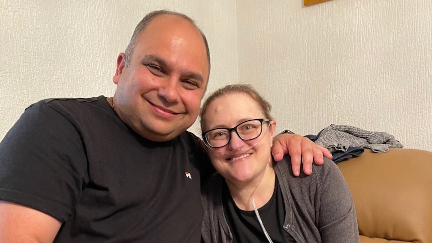 a man smiling and hugging a woman wearing glasses and a breathing tube 