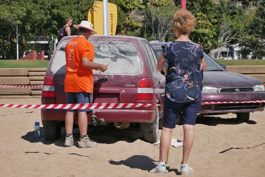 A woman watches artist Scott Wade painting in dirt on a car window.