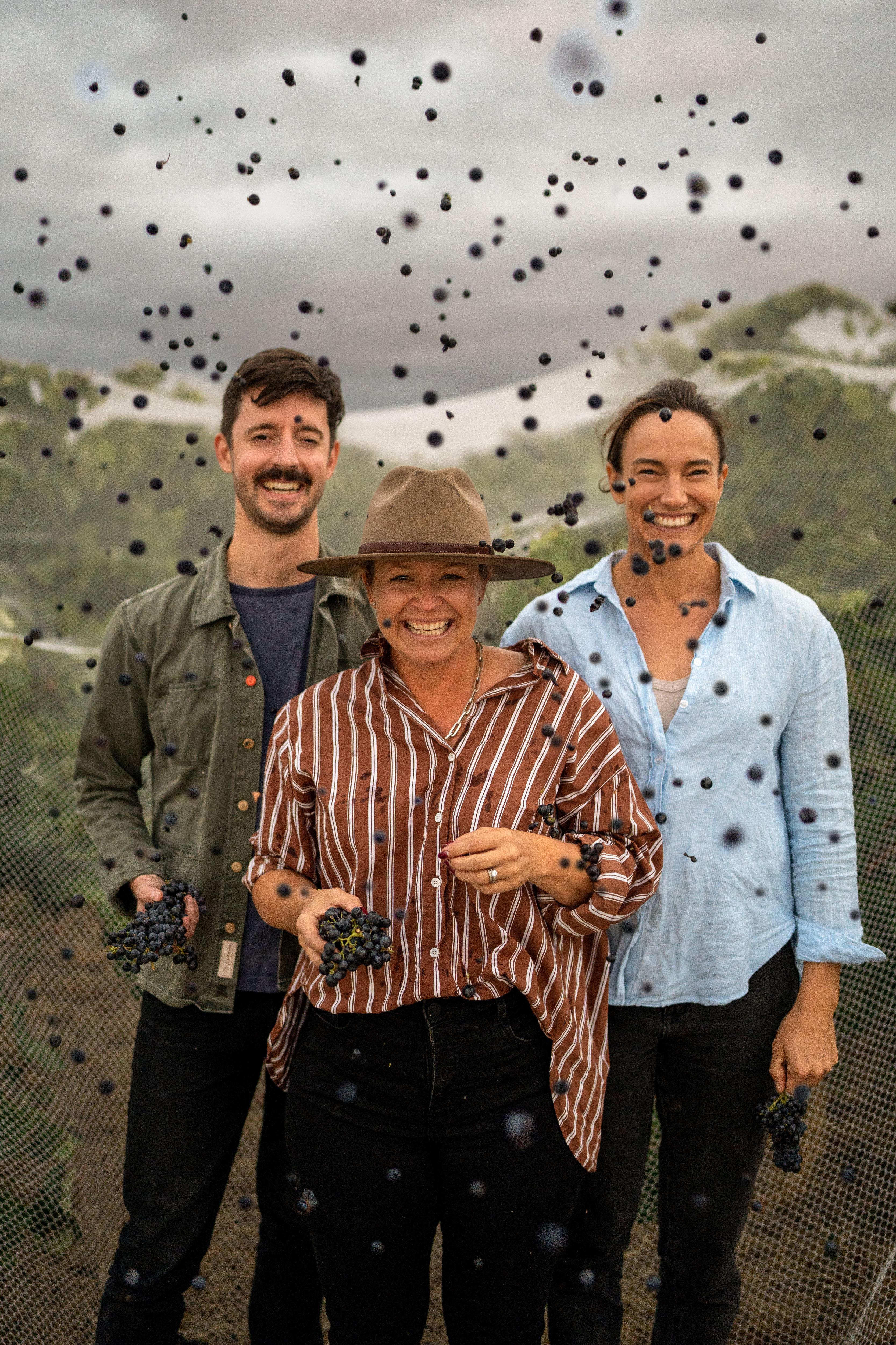 A man and two women smile with grapes surrounding them in the air.