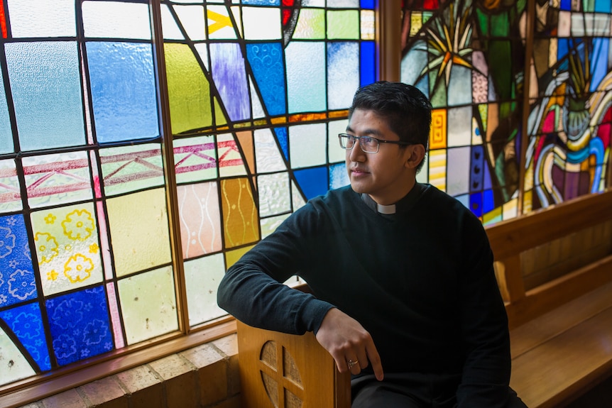A priest dressed in black with a white collar, sits on a pew in front of a wall of stained glass.