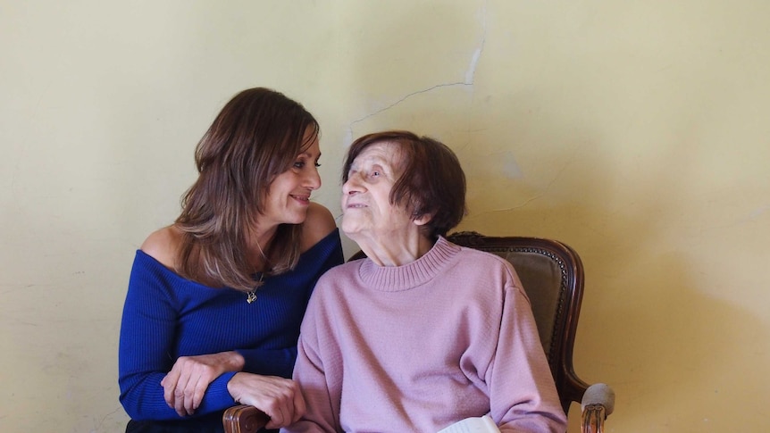 A middle aged woman wearing blue sitting next to her elderly mother wearing pink and gazing into her eyes