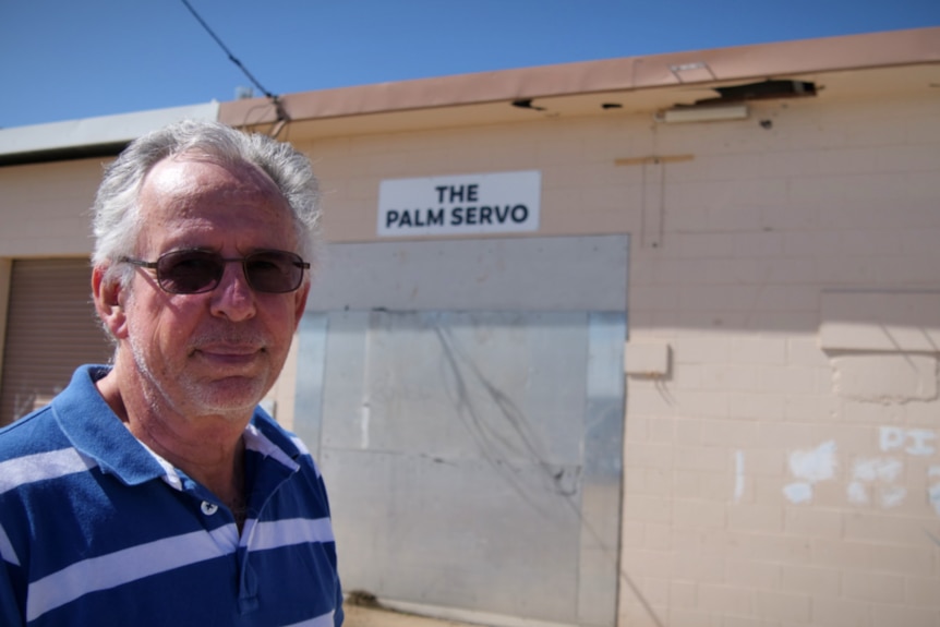 A grey haired man in a blue and white shirt stands out the front of a building with the words the palm servo on it