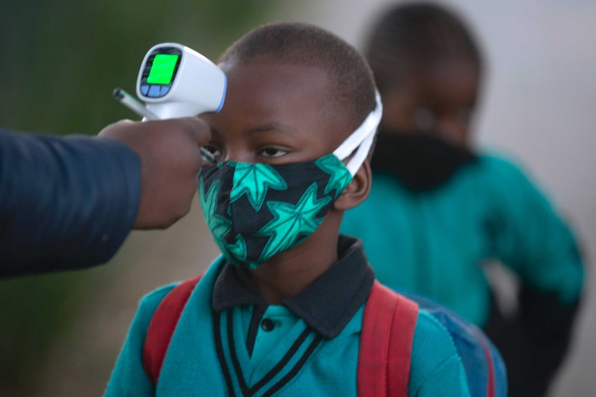 A small African boy wearing green stands silently as a white temperature gun checks him for fever.