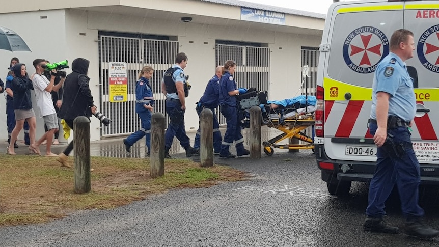 A group of media watch on as paramedics cart a man on a bed towards an ambulance.