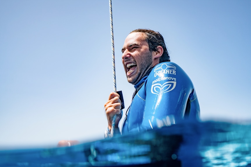A man in sea water wearing a blue wetsuit laughs.