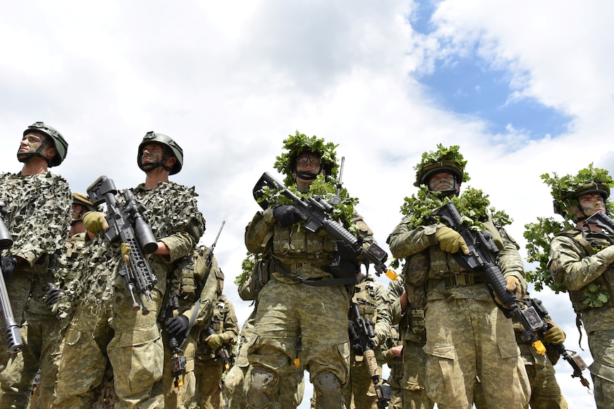A line of soldiers wearing camouflage hold weapons, image taken from low angle looking up.