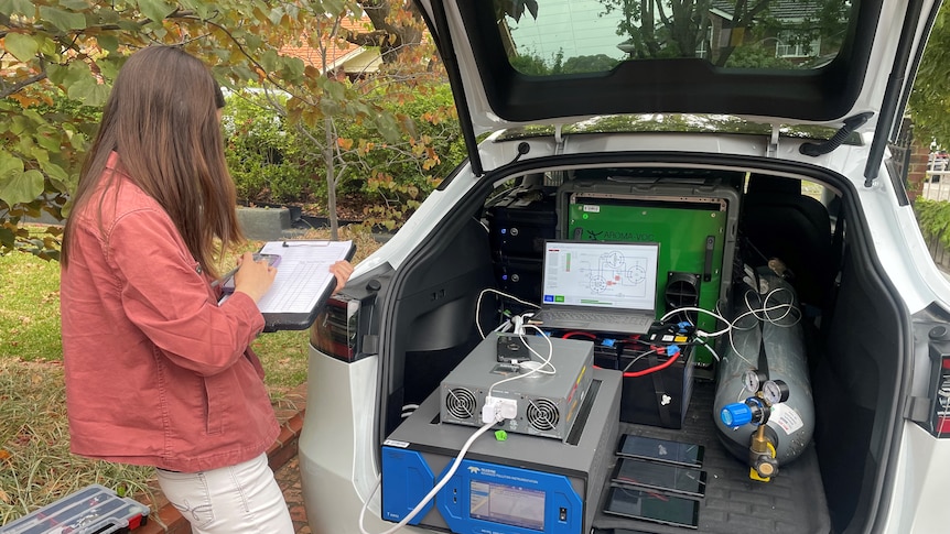 A woman writes on a clipboard, standing in front of a Tesla car boot filled with machinery.