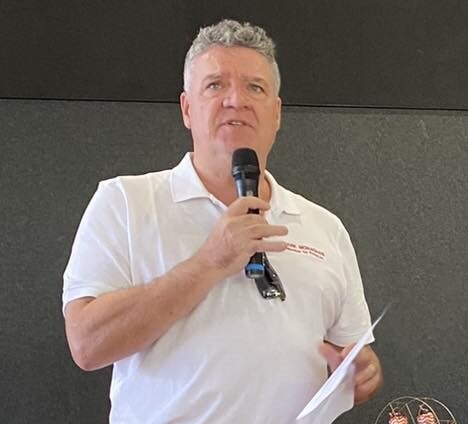 A white man with short grey hair speaking into a microphone at an indoor event