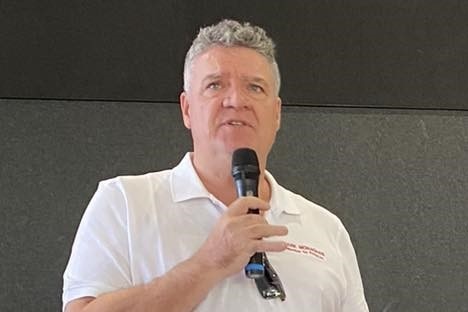A white man with short grey hair speaking into a microphone at an indoor event