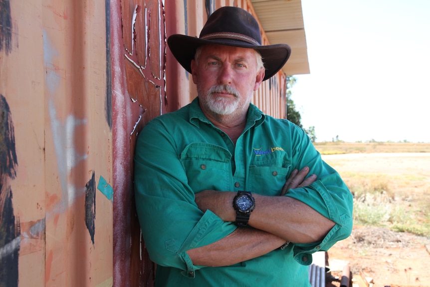 A man with a white beard leans on a fence wearing a green shirt arms crossed frowning