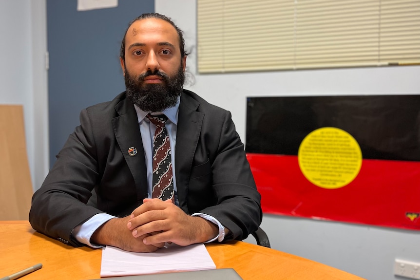 A bearded man in a business suit at a desk with and Aboriginal flag in the background.