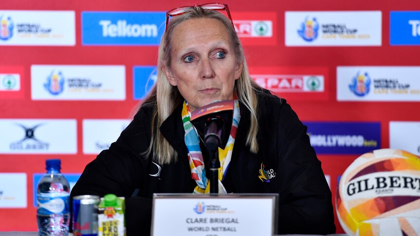 A world netball executive sits at a desk at a press conference and looks toward the media as she is asked a question. 