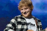 A mid shot of a smiling Ed Sheeran on stage playing his guitar against a blue backdrop at Perth Stadium.