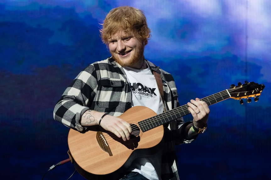 A mid shot of a smiling Ed Sheeran on stage playing his guitar against a blue backdrop at Perth Stadium.