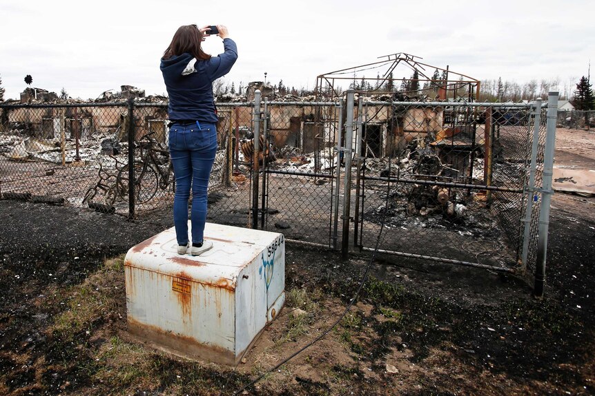 A woman takes a photograph over a fence at the burned skeleton of a house.