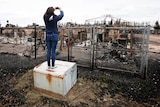 A woman takes a photograph over a fence at the burned skeleton of a house.