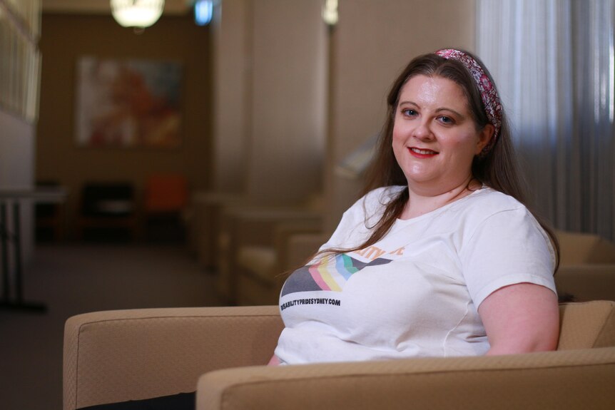 A young woman sitting in a lounge chair. She is wearing a white t-shirt about disability pride