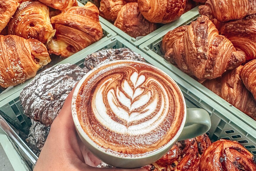 A hand holds a cup of coffee with fancy latte art as pastries wait for costumers in the background