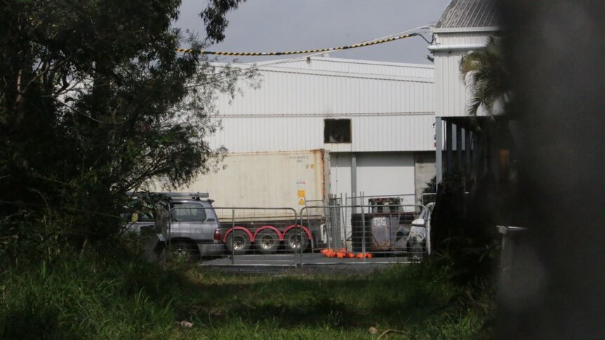 A big white factory shed surrounded by fencing and trees with a truck next to it.