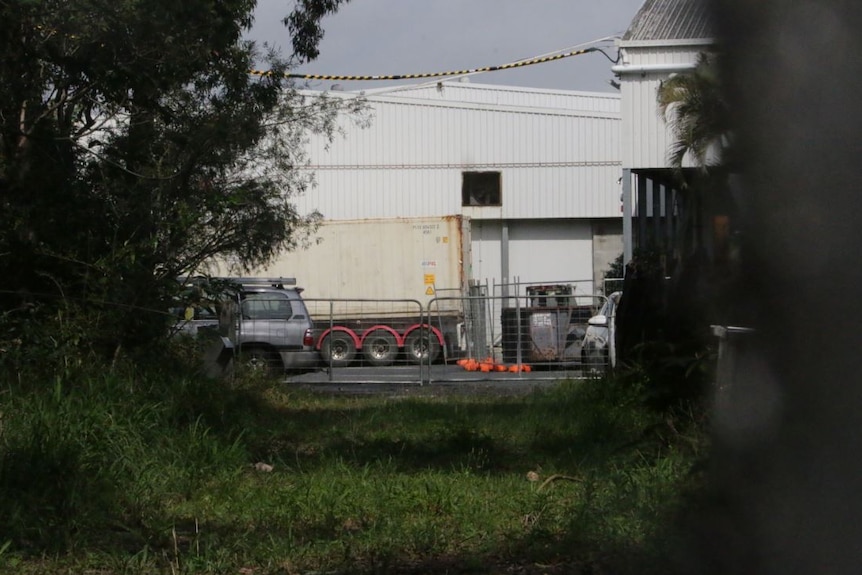 A big white factory shed surrounded by fencing and trees with a truck next to it.