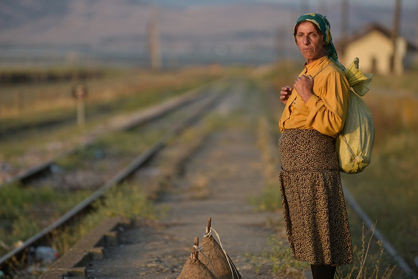 An older woman wearing green headscarf, yellow top and carry yellow sack on back stands with by train tracks in the countryside.