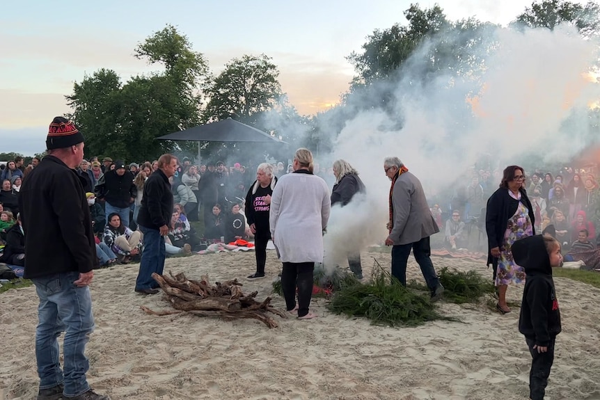 People walk through the smoke coming from burning branches on the sand in front of a large crowd
