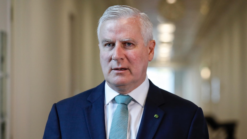 A man with grey hair wearing a navy suit and aqua tie standing in front of a long white corridor