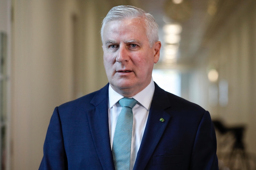 A man with grey hair wearing a navy suit and aqua tie standing in front of a long white corridor