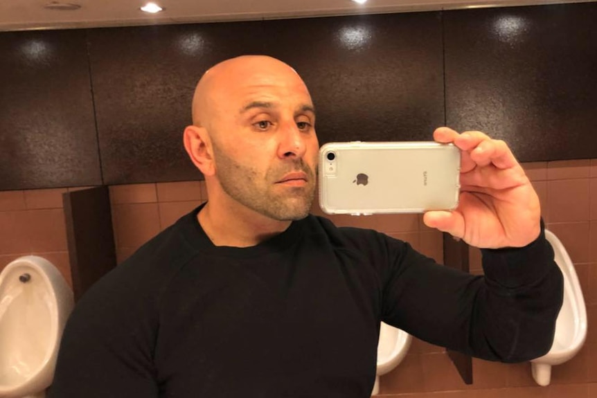 A bald man with a light beard taking a selfie in a mirror using a phone