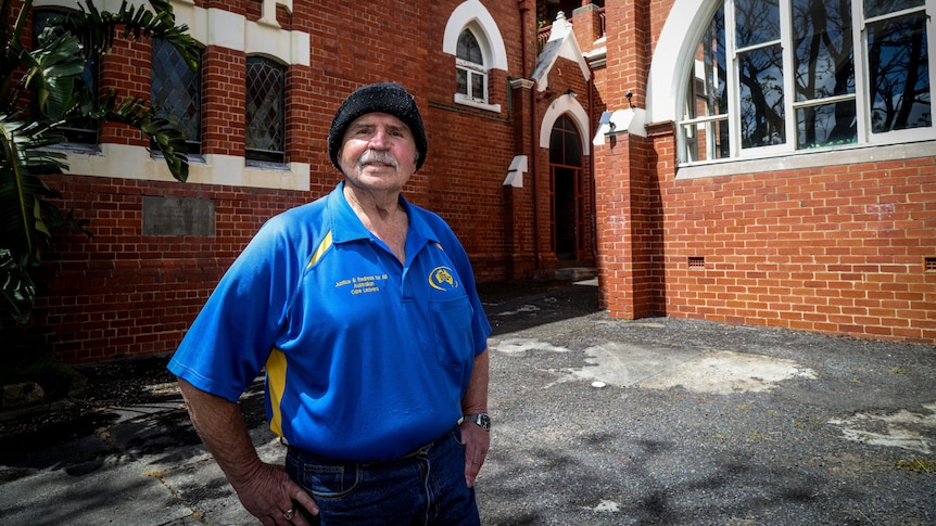 A man wearing a black beanie and a bright blue shirt stands in front of a red brick building.