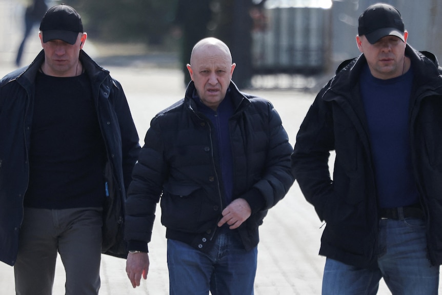 A bald man walks between two beefy security guards in caps