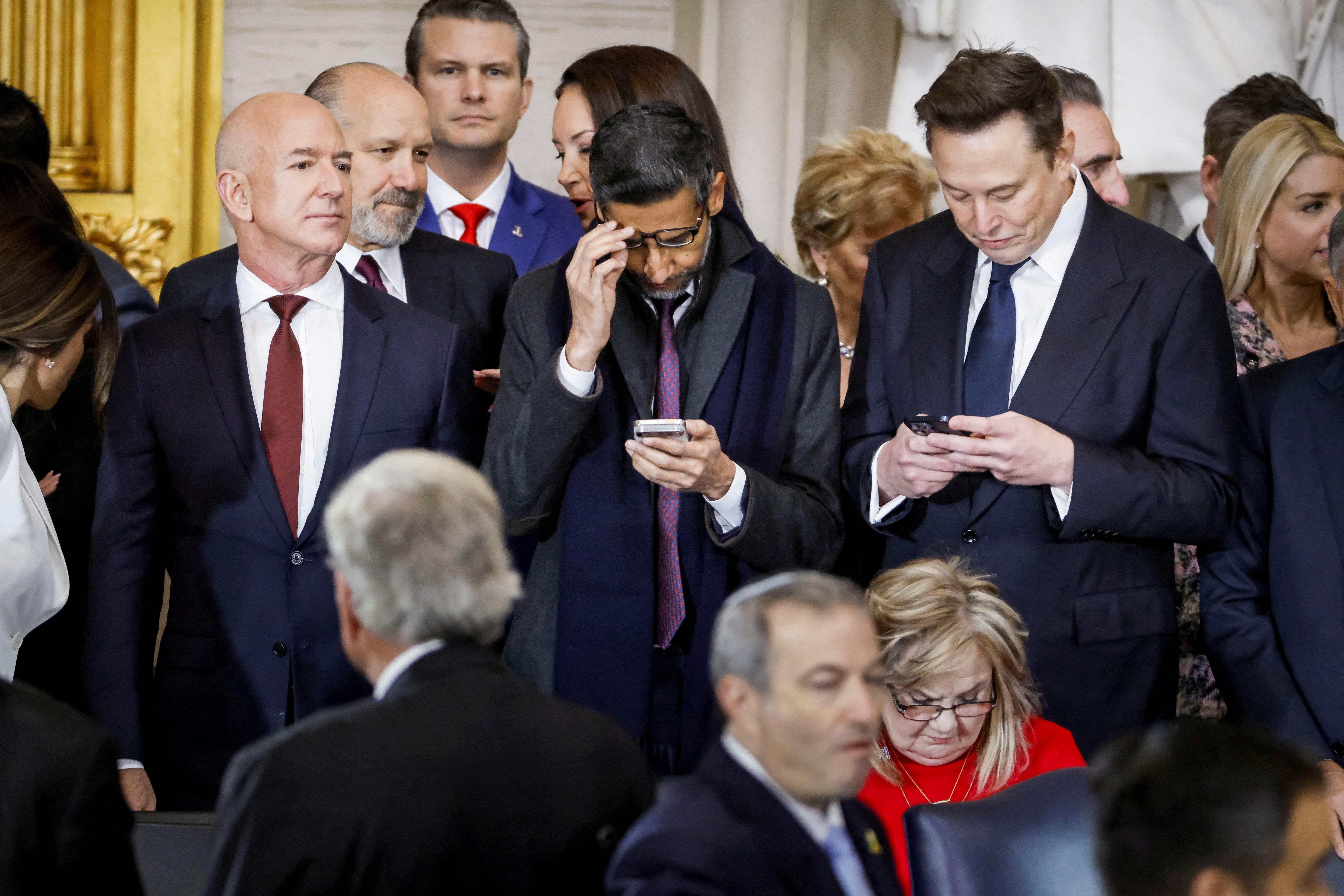 Three men wearing suits at an official event. Two of them are looking at their phones in their hands.