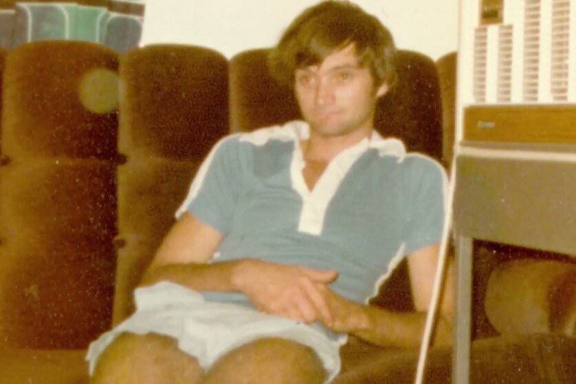 A young Gregory Smith sitting on a brown coach, brown hair, blue shirt, light shorts.