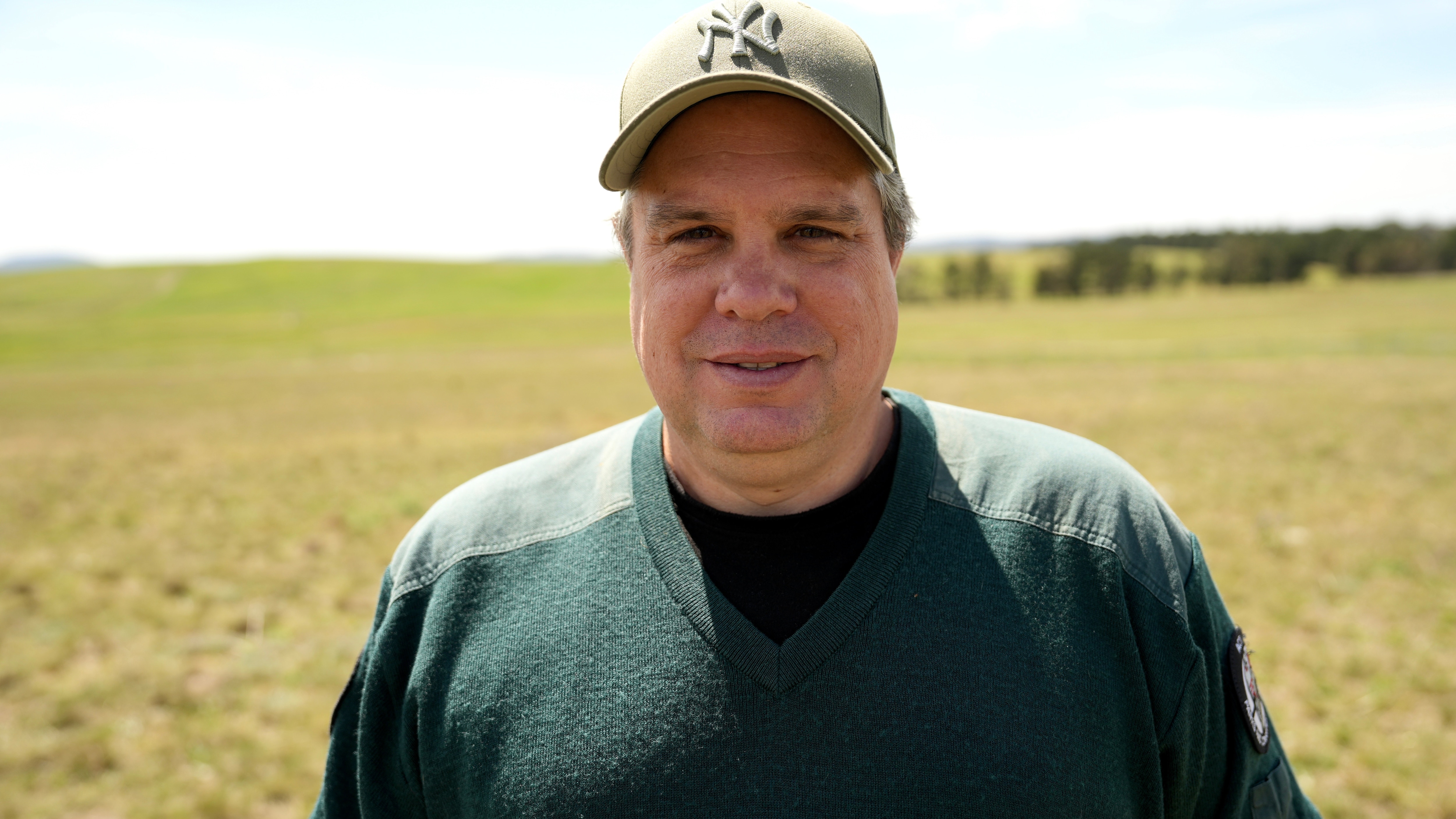 A man wearing a dark green sweater and beige cap stands in a paddock smiling lightly.