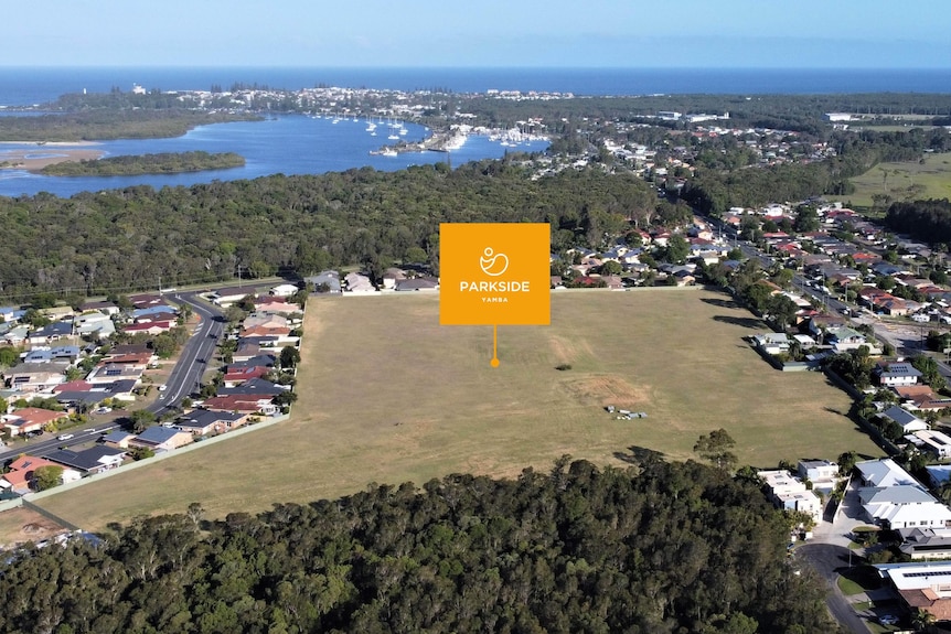An aerial view of a large vacant block surrounded by homes and a nature reserve with a river mouth and ocean in the distance.