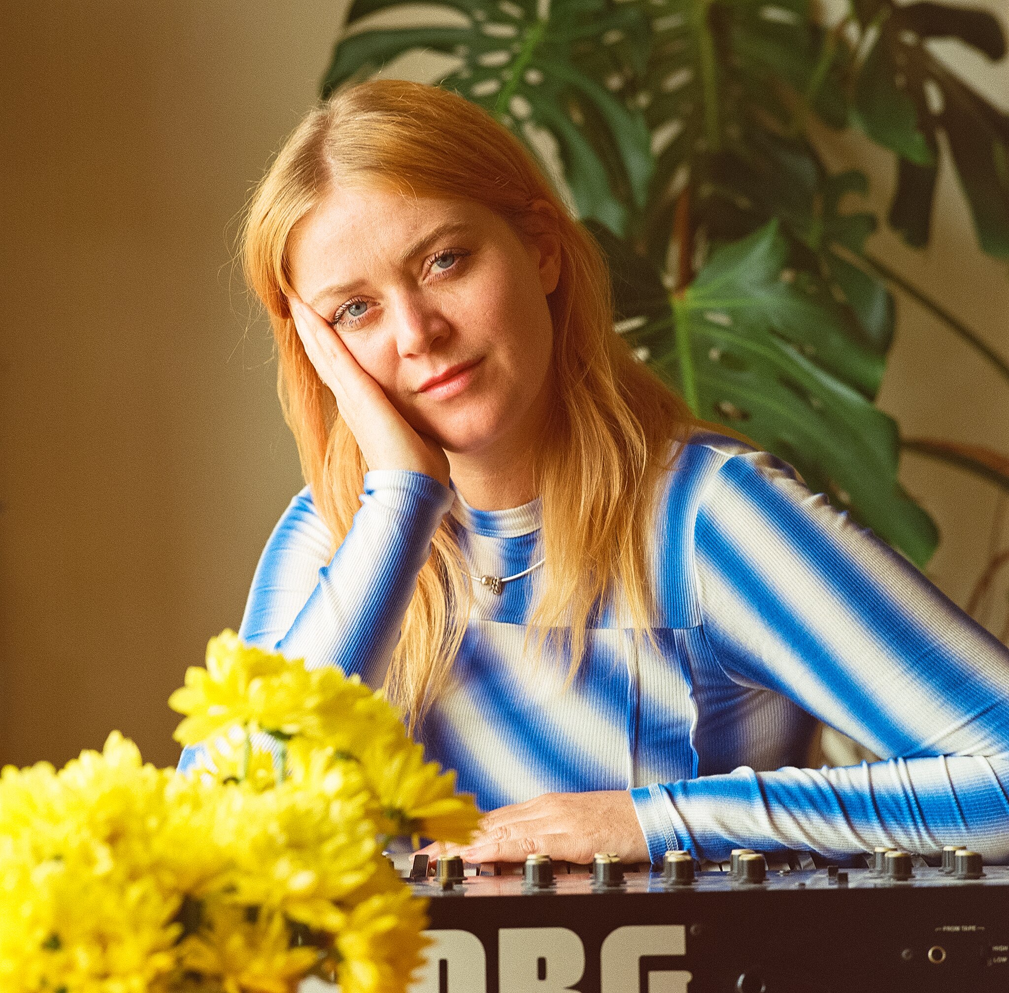 Ella Thompson sitting in front of a mixing desk with flowers in the foreground. She is wearing a long sleeve shirt.